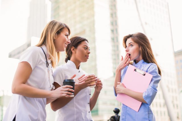 Three young nurses stand with cups of coffee and folder in hands and discuss about something with surprise during coffee break, outdoors