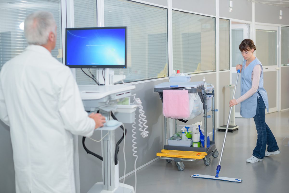 Woman cleaning hospital corridor, doctor using computer