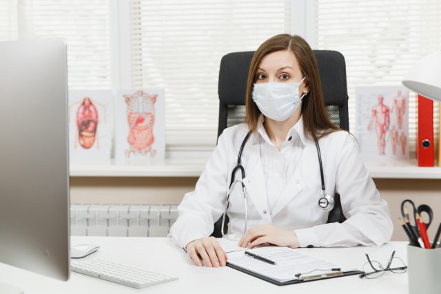 Young female doctor sitting at desk, working on modern computer with medical documents in light office in hospital. Woman in medical gown, face mask in consulting room. Healthcare, medicine concept.