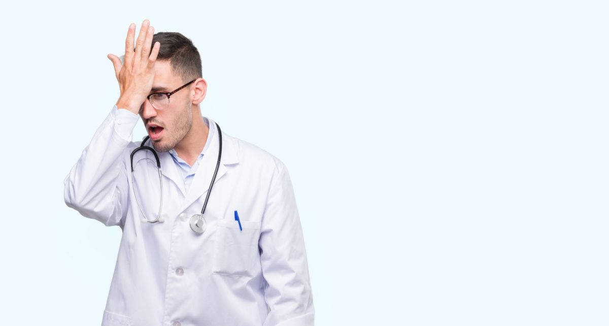 Handsome young doctor man surprised with hand on head for mistake, remember error. Forgot, bad memory concept.